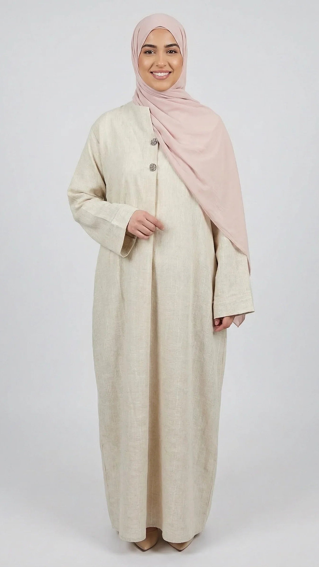 Femme souriante portant une abaya beige élégante Muneera et un hijab rose, mode modeste moderne.