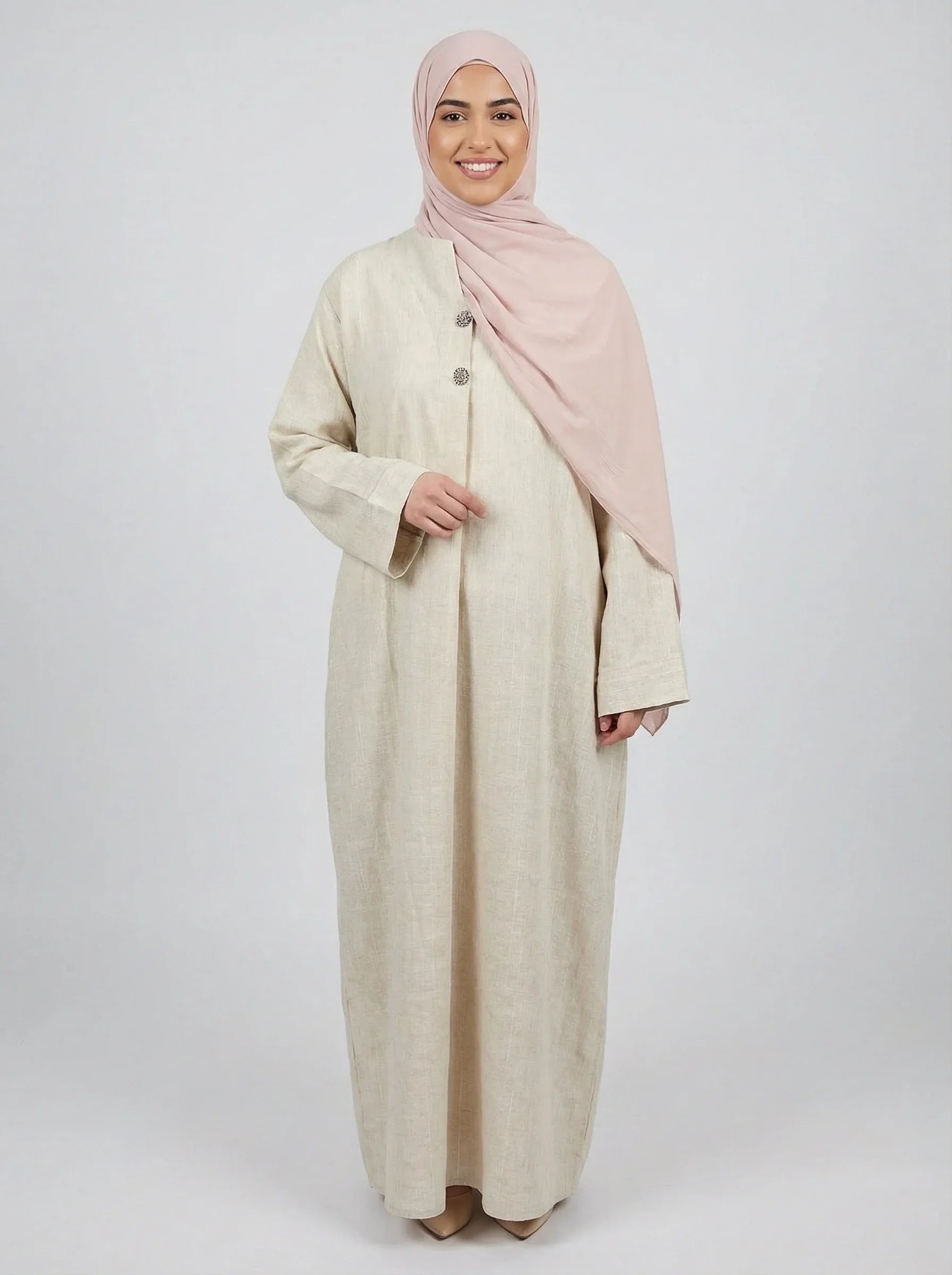 Femme souriante portant une abaya beige élégante Muneera et un hijab rose, mode modeste moderne.
